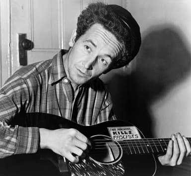 Woody Guthrie: half-length portrait, seated, facing front, playing a guitar that has a sticker attached reading: This Machine Kills Fascists